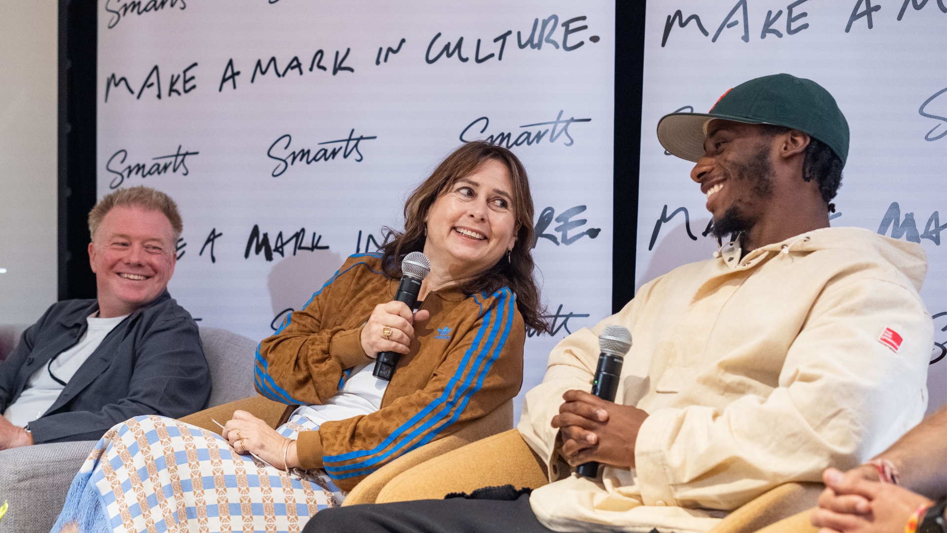 Three people holding microphones are engaged in a lively discussion while seated on stage with "MAKE A MARK IN CULTURE" and “SMARTS” logo banners in the background.