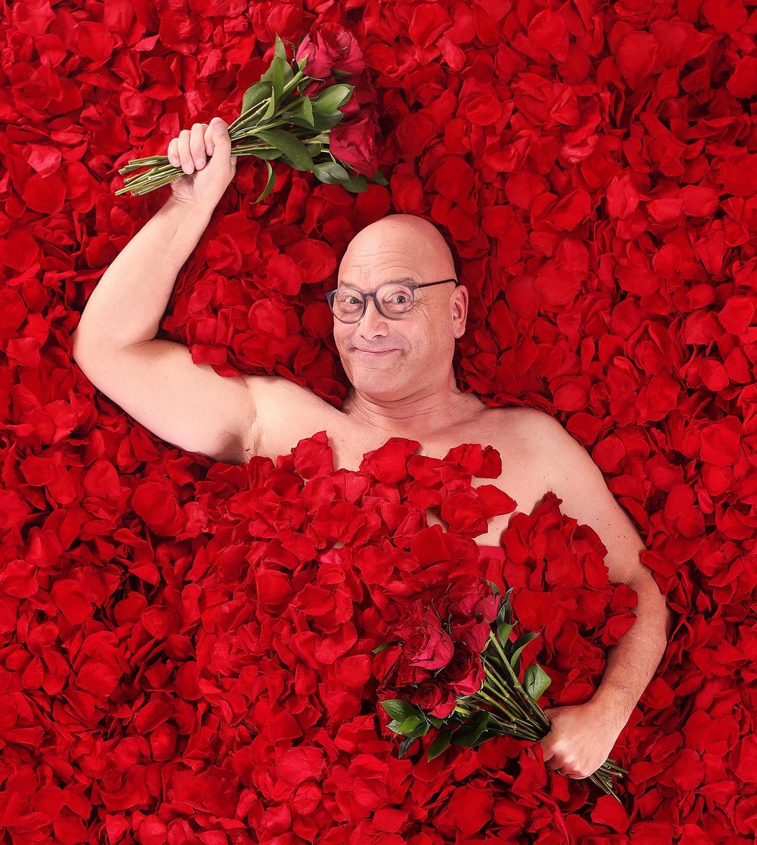 Greg Wallace, a white man wearing glasses and lying on a bed of red roses covering his topless chest. Holding two bunches of roses in both hands. 