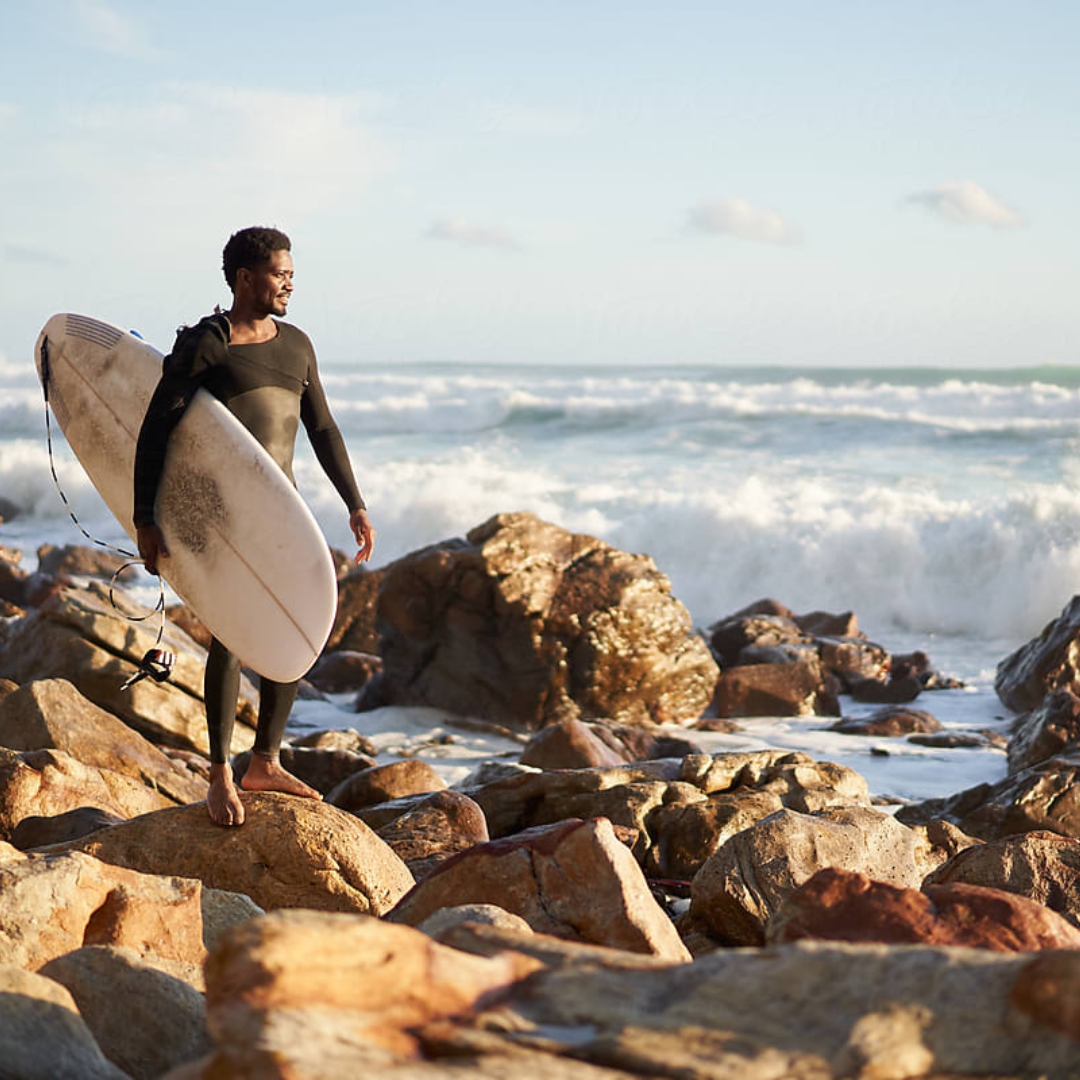 Man standing on rocks at the beach wearing wetsuit and holding surfboard
